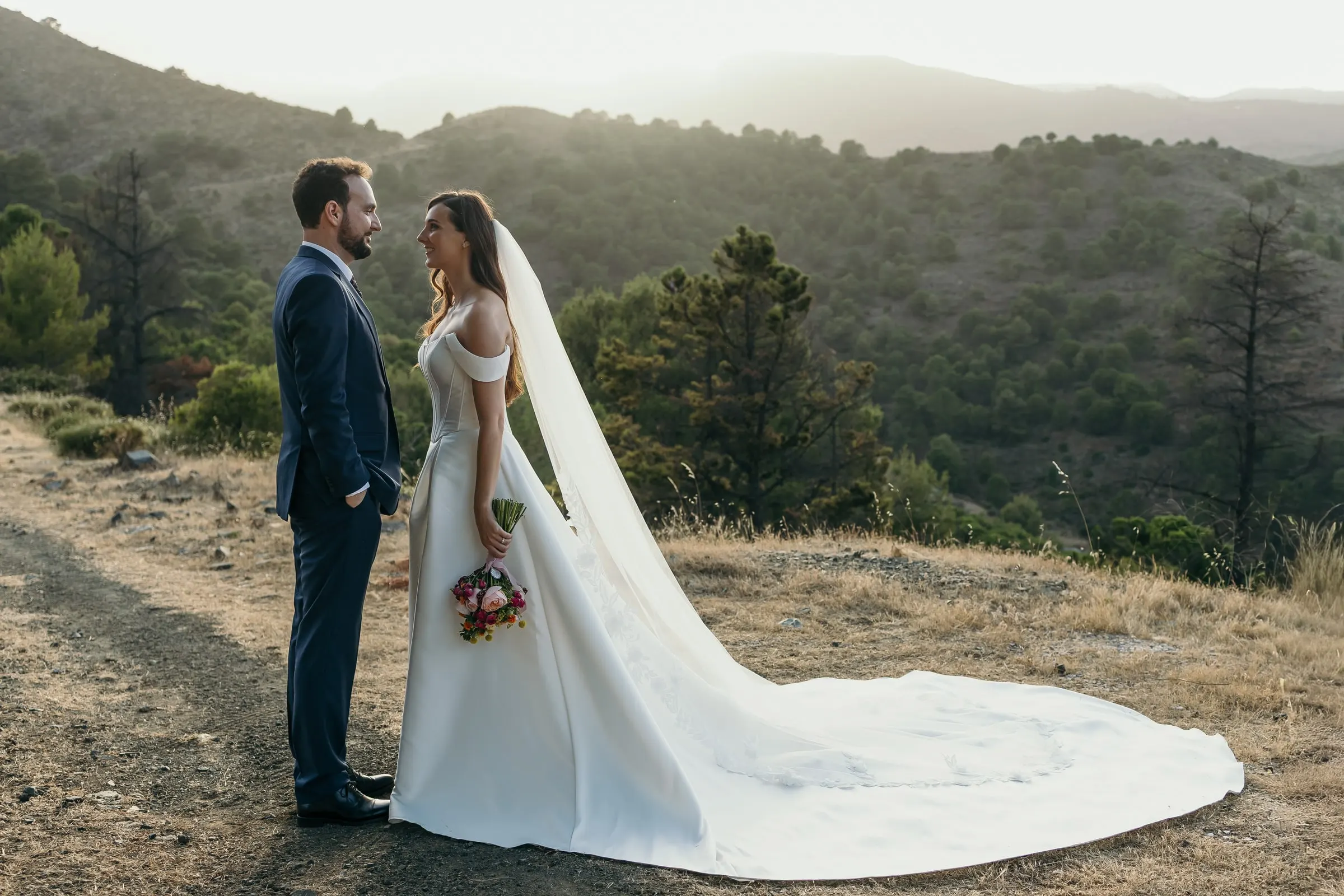 Los novios frente a frente en un camino en lo alto de la sierra con capas de montañas detrás