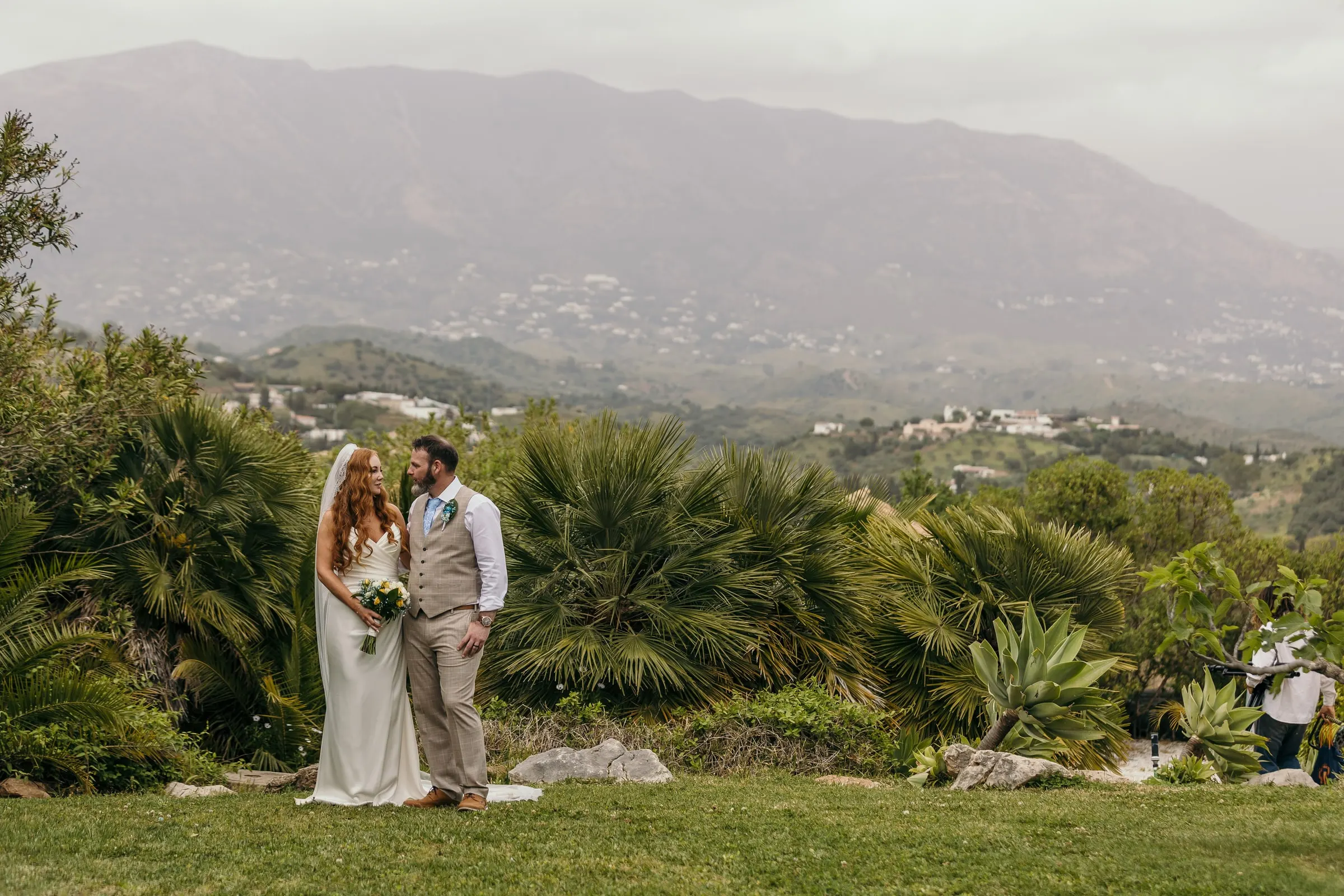 Los novios entre palmitos en el césped de Hacienda San José, con la sierra de Mijas y sus pueblos blancos detrás