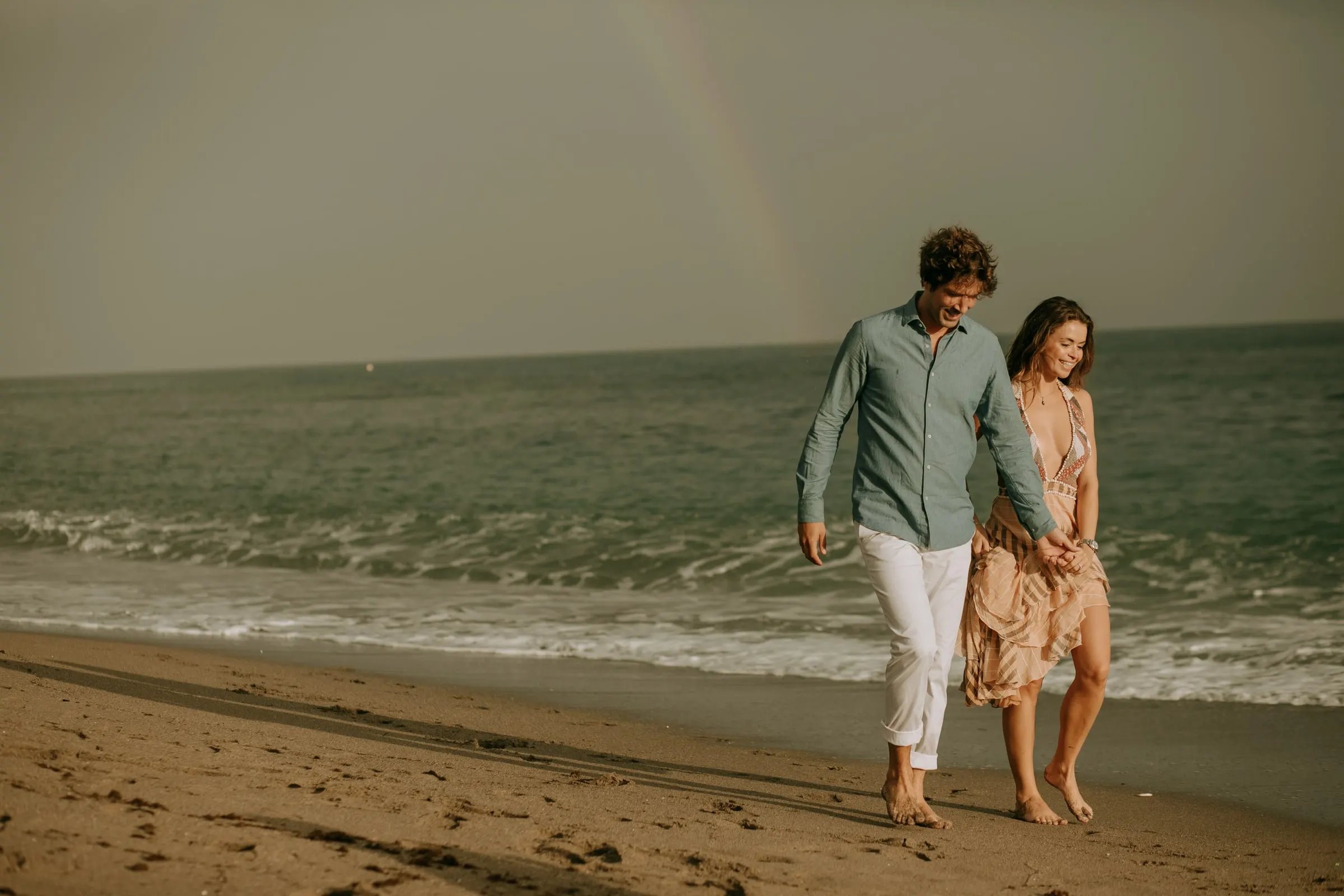 Lauren y Arnaud caminan descalzos por la orilla con la luz suave del atardecer, sesión preboda en la playa de Sotogrande